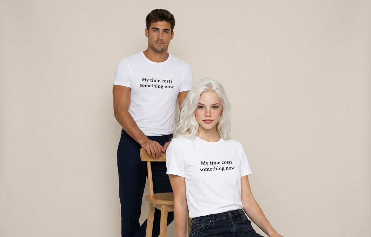 A man and a woman modeling matching white graphic t-shirts with the black printed text 'My time costs something now'. The man stands behind a wooden stool wearing dark pants, while the woman sits in front of it wearing dark jeans.