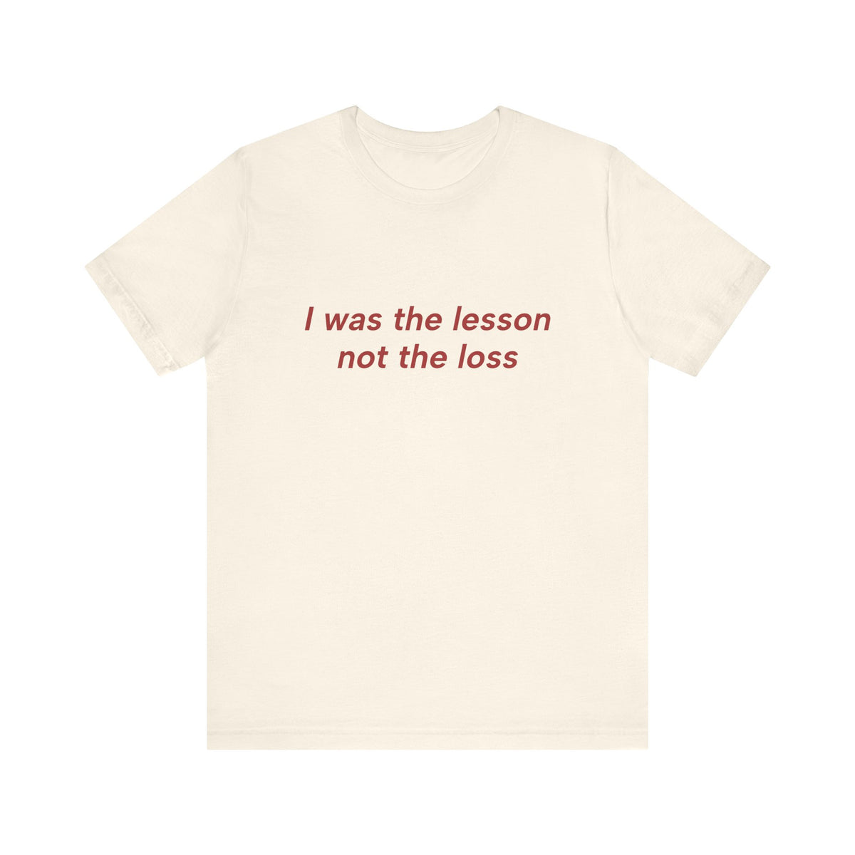 An off-white short-sleeve crewneck t-shirt with the phrase 'I was the lesson not the loss' printed in red serif text across the chest, displayed on a solid white background.
