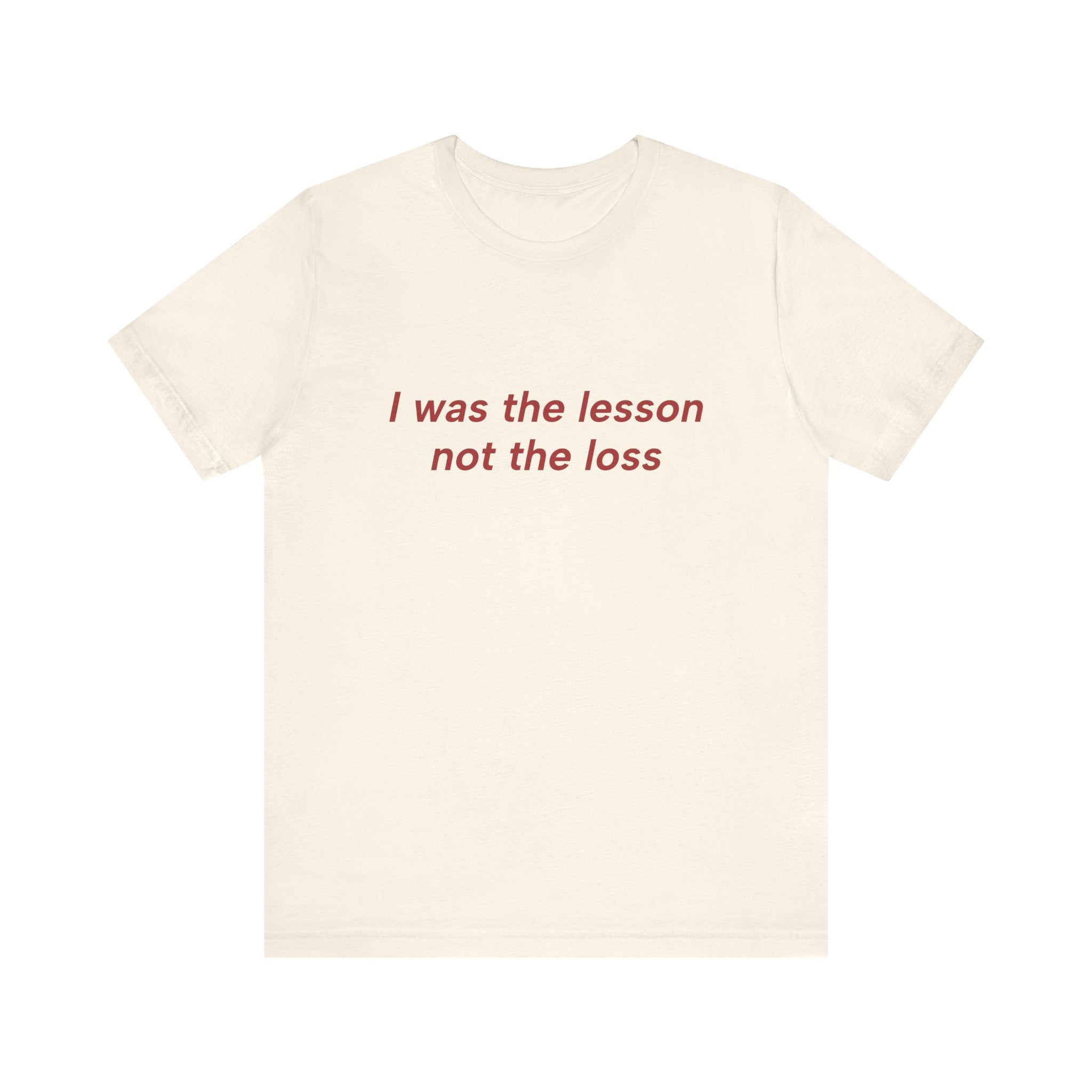 An off-white short-sleeve crewneck t-shirt with the phrase 'I was the lesson not the loss' printed in red serif text across the chest, displayed on a solid white background.