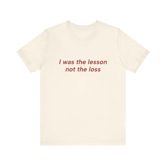 An off-white short-sleeve crewneck t-shirt with the phrase 'I was the lesson not the loss' printed in red serif text across the chest, displayed on a solid white background.