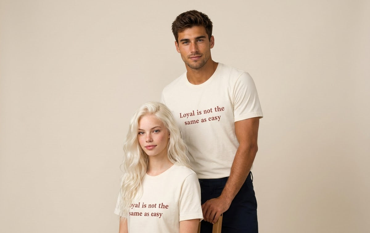 Two models wearing matching cream-colored graphic t-shirts with the text 'Loyal is not the same as easy' printed in a reddish-brown serif font, posed against a solid beige studio background.
