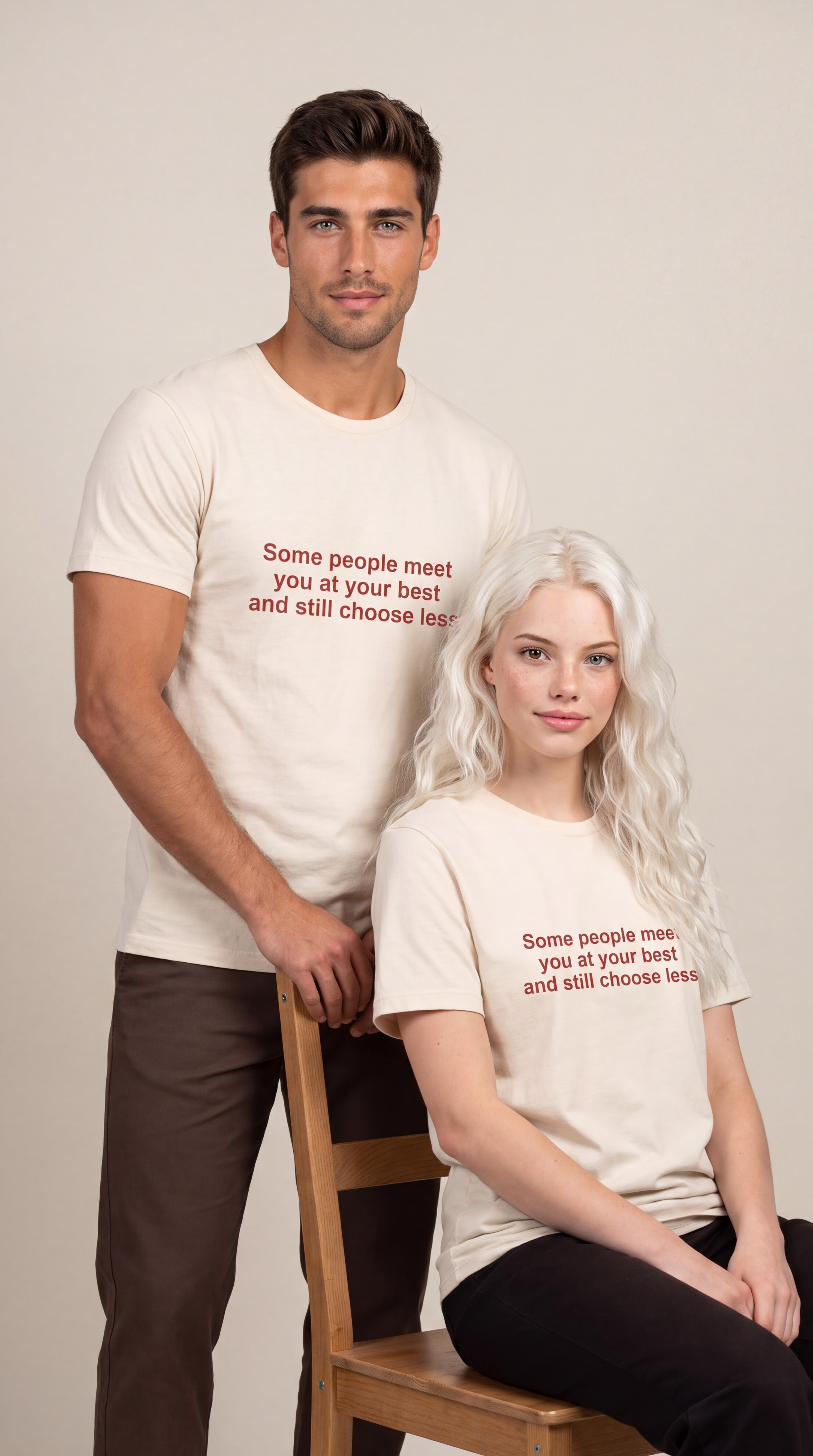 A man and a woman modeling matching cream-colored t-shirts with red text that reads 'Some people meet you at your best and still choose less.' The man stands behind the woman, who is seated on a wooden chair, against a plain beige studio background.