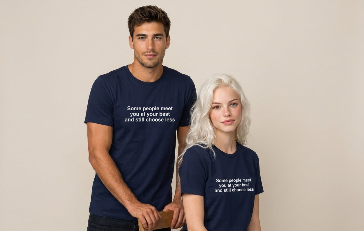 A man and a woman modeling matching navy blue t-shirts with white text that reads, 'Some people meet you at your best and still choose less.' The man stands holding a wooden chair, and the woman sits beside him against a plain beige studio background.