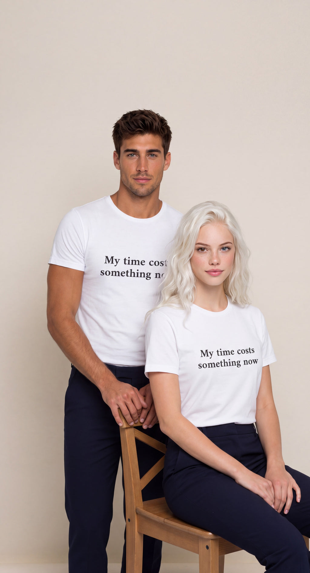 A man and a woman modeling matching white graphic t-shirts with the black printed text 'My time costs something now.' The man stands behind the woman, who is seated on a wooden chair, against a plain beige studio background.

