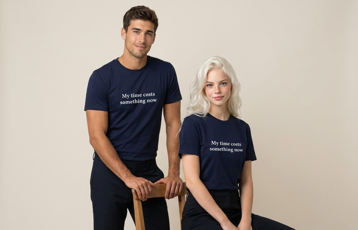 A man and a woman modeling matching navy blue t-shirts with the white printed text 'My time costs something now'. The man stands on a wooden chair while the woman sits on the floor against a plain beige studio background.