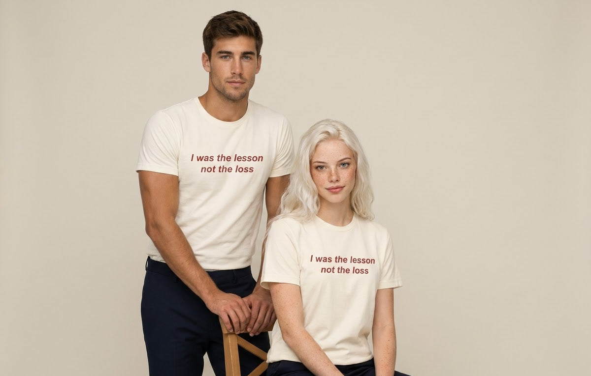 A man and a woman modeling matching white graphic t-shirts with the printed text 'I was the lesson not the loss' in red. The man stands on a wooden chair, and the woman sits beside him against a solid light beige background.