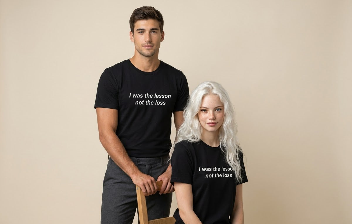 A man and a woman modeling matching black graphic t-shirts with the white printed text 'I was the lesson not the loss' against a solid beige studio background.