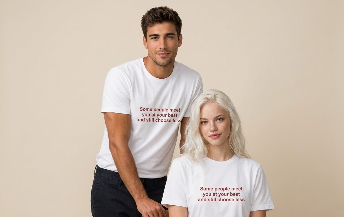 A man and a woman wearing matching white graphic t-shirts with the text 'Some people meet you at your best and still choose less' printed in a dark red serif font, posed against a solid beige background.