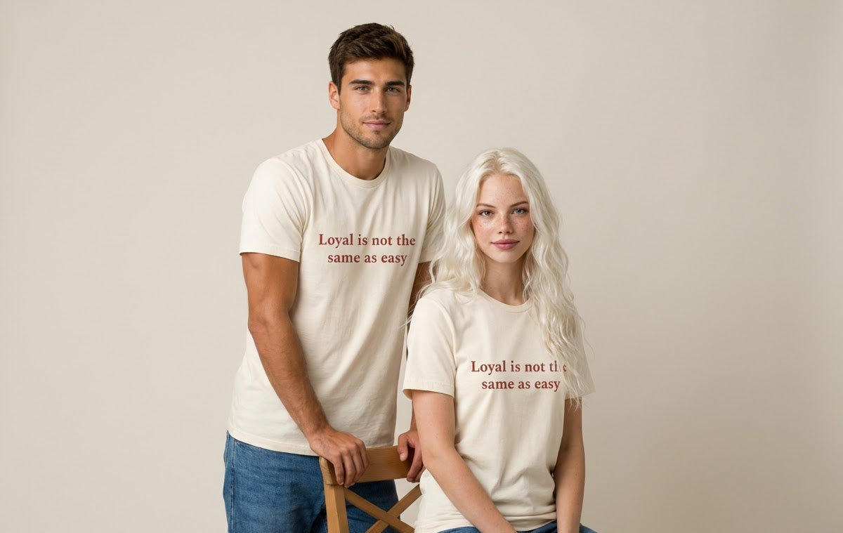 A man and a woman modeling matching cream-colored t-shirts with the text 'Loyal is not the same as easy' printed in red on the chest, posed against a solid light beige background.