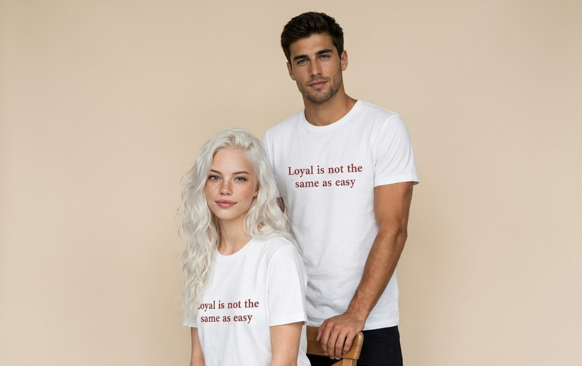 A man and a woman wearing matching white graphic t-shirts with the text 'Loyal is not the same as easy' printed in red, posed against a solid beige studio background.