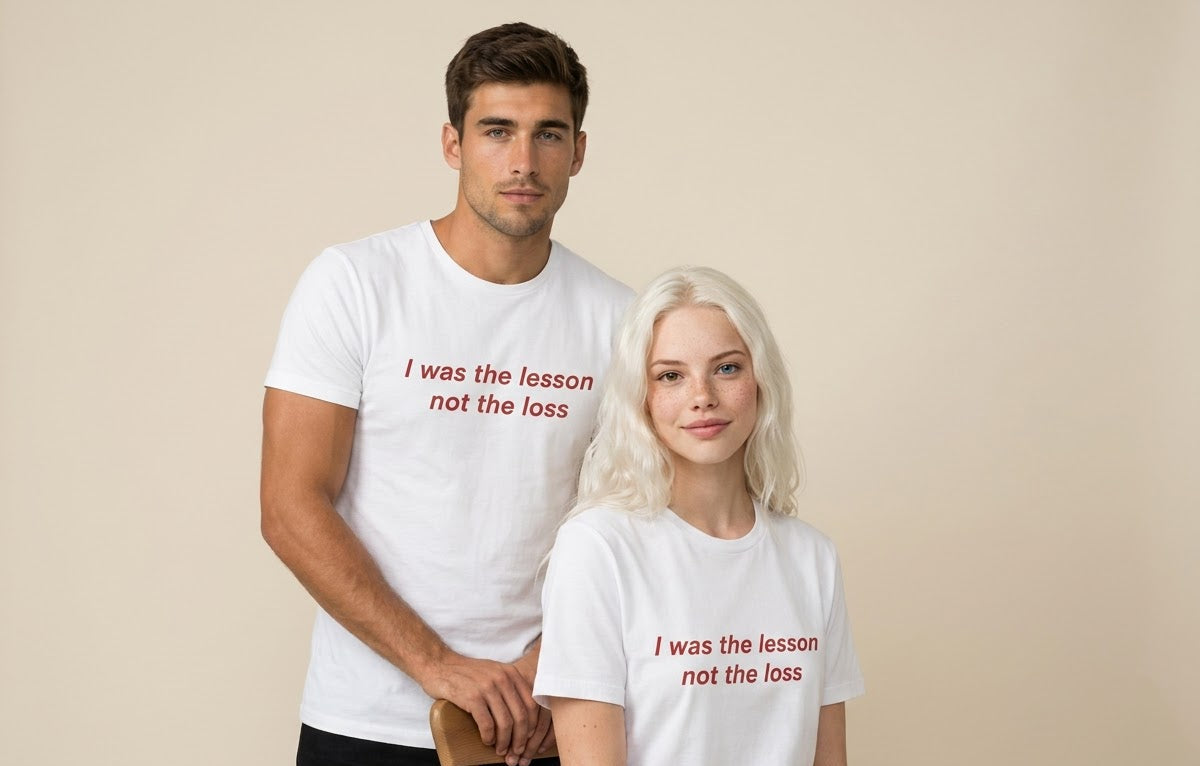 A man and a woman wearing matching white graphic t-shirts with the red printed text 'I was the lesson not the loss', posed against a solid beige background.