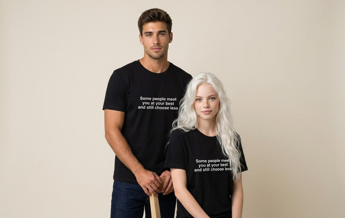 A man and a woman modeling matching black graphic t-shirts with white text that reads 'Some people meet you at your best and still choose less'. The man stands behind the woman, who is seated on a light wood chair, against a plain beige studio background.